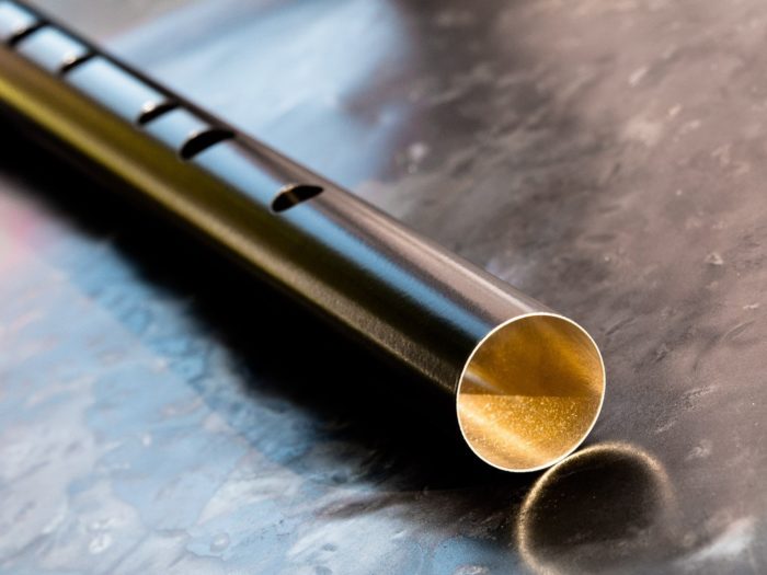 Black and gold, brass bodied, low d tin whistle on a shinny metallic background. Angle of image clearly showing polished brass interior of whistle body. Five contoured finger holes can also be seen.